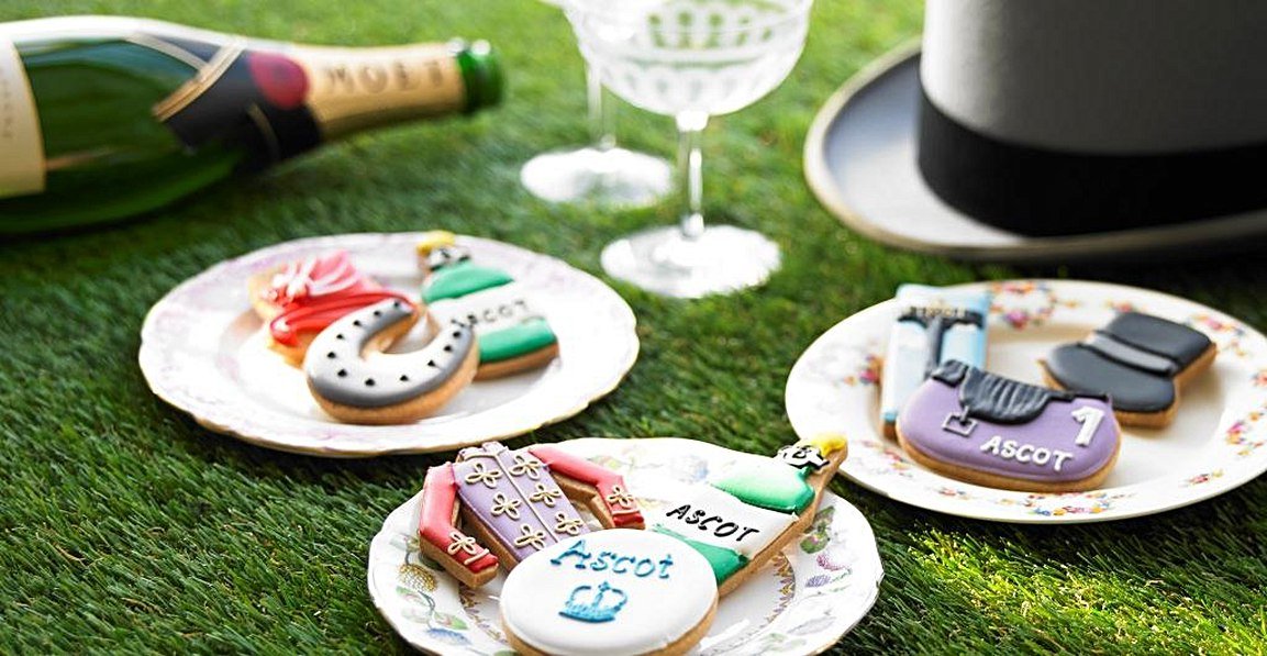 Ascot-themed biscuit collection on grass, featuring royal silks, horse shoe, and Ladies' Day hat by Biscuiteers for personalized gifts.